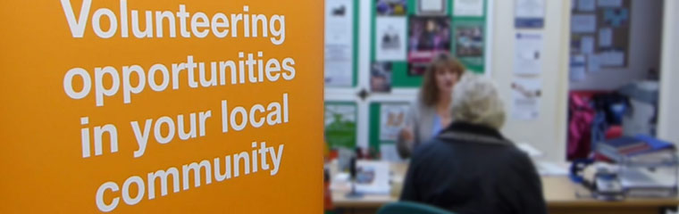 A sign saying volunteering opportunities in your local community next to a woman sat at a desk talking to other woman.