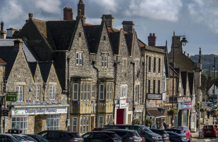 Row of shops along the high street including Chipping Sodbury Pharmacy. Cars are parked in front of the shops.