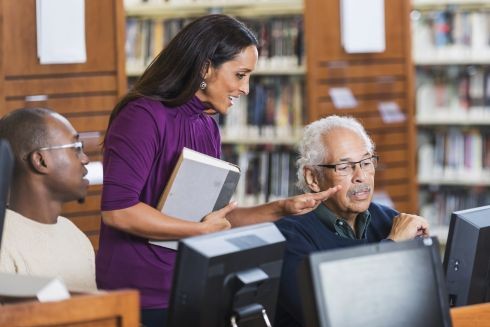 An older man sitting alongside a man in his 30s at computers in a community venue receiving instruction from a woman