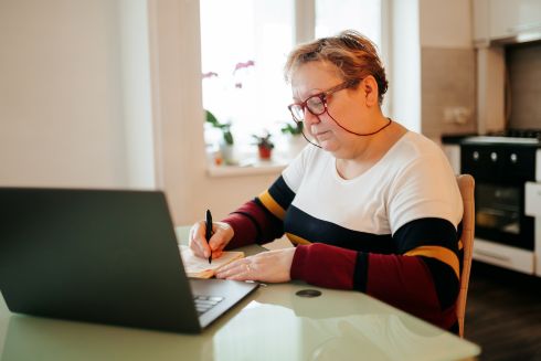 Middle aged woman using a laptop and taking notes with a pen and notepad sitting at a kitchen table